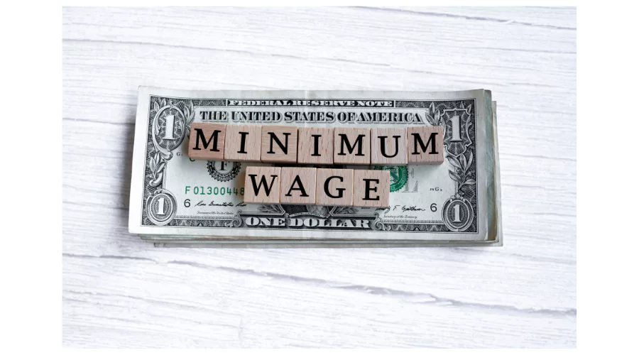 A stack of one-dollar bills with 'MINIMUM WAGE' stamped across them on a white wooden background, indicating discussions about the 'minimum wage in Florida' and its economic implications.