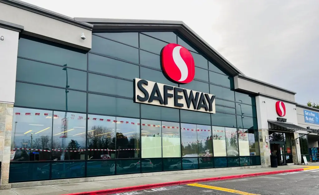 Exterior view of a Safeway store with prominent signage, reflecting the busy street on its large glass facade, with text on the window promoting a free flu shot, indicative of the various services offered like Safeway Money Orders.