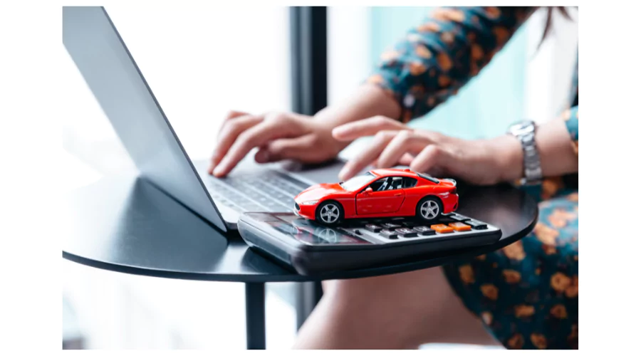A person's hands typing on a laptop next to a small red toy car placed on a calculator, symbolizing the online management or application for an auto loan or vehicle financing.