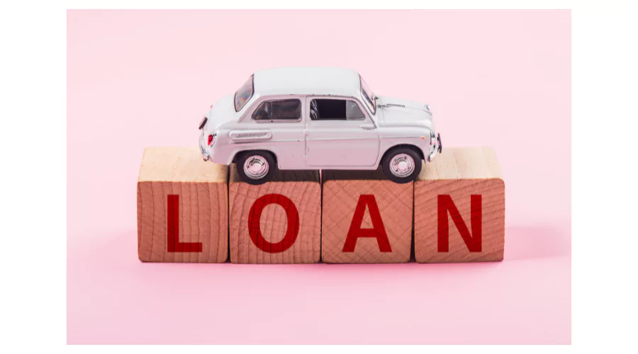 A miniature white car model placed atop wooden blocks spelling out the word 'LOAN' in red capital letters, against a soft pink background. This represents the concept of an auto equity loan where a vehicle is used as collateral.