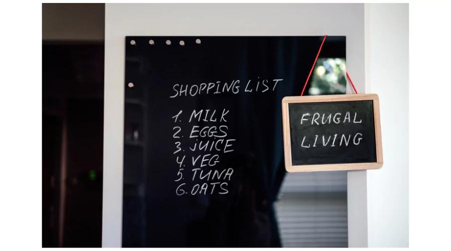 A shopping list written on a blackboard displaying items like milk, eggs, juice, vegetables, tuna, and oats, next to a small chalkboard hanging on the wall with 'Frugal Living' written on it, symbolizing the practice of budgeting and mindful spending for a cost-effective lifestyle.