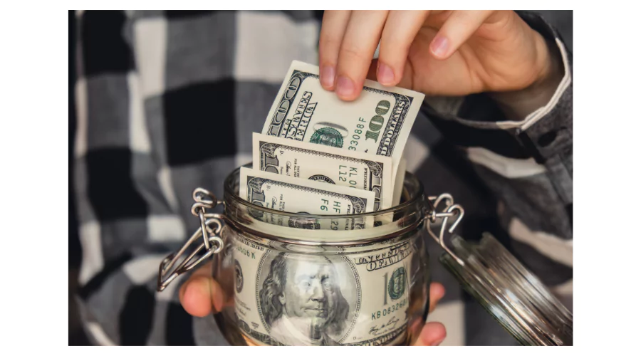 A person's hand placing folded dollar bills into a glass jar, representing a simple yet effective 'money hack' for saving and accumulating wealth.