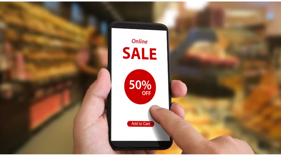 A hand holding a smartphone with an 'Online Sale 50% OFF' advertisement on the screen, set against the blurred backdrop of a shopping aisle, illustrating how the Flipp app revolutionizes savings for smart shoppers.