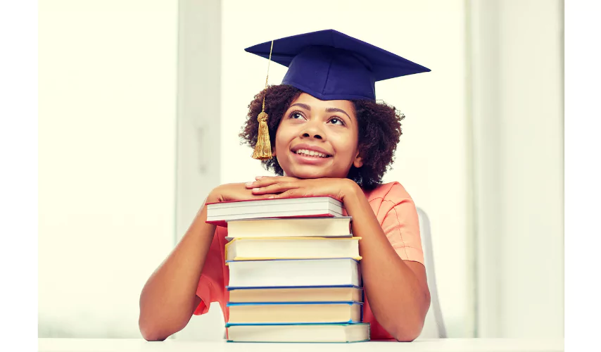 Young African American girl wearing a graduation cap, resting her chin on a stack of colorful textbooks, looking upward with a hopeful expression, symbolizing academic success through PrepScholar.